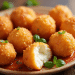 A plate of golden, crispy Cottage Cheese Balls garnished with fresh herbs sits on a wooden table. One ball is bitten open, revealing a creamy white filling inside. A reddish sauce is spread beneath the cheese balls.
