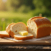 A loaf of bread is partially sliced on a wooden cutting board outdoors, with a couple of slices topped with pats of butter—a perfect outdoor boys bread recipe moment set against a sunny, blurred backdrop of green grass.
