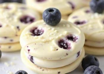 Round blueberry cream cookies are stacked on a white surface, with fresh blueberries scattered around and on top. These white chocolate blueberry Oreos have visible blueberry pieces and a creamy texture.