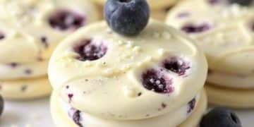 Round blueberry cream cookies are stacked on a white surface, with fresh blueberries scattered around and on top. These white chocolate blueberry Oreos have visible blueberry pieces and a creamy texture.