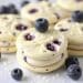 Round blueberry cream cookies are stacked on a white surface, with fresh blueberries scattered around and on top. These white chocolate blueberry Oreos have visible blueberry pieces and a creamy texture.