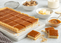 A rectangular caramel slice tart, made with an easy caramel slice recipe, is cut into squares on a white tray. Nearby are bowls of caramel fudge, butter, cream, and a glass jar of caramel sauce on a light background.
