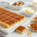 A rectangular caramel slice tart, made with an easy caramel slice recipe, is cut into squares on a white tray. Nearby are bowls of caramel fudge, butter, cream, and a glass jar of caramel sauce on a light background.