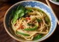 A bowl of noodle soup with green vegetables and herbs, served in a patterned blue and white bowl on a wooden table with chopsticks on the edge, evokes the comforting flavors of a classic phat si-io recipe.