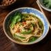 A bowl of noodle soup with green vegetables and herbs, served in a patterned blue and white bowl on a wooden table with chopsticks on the edge, evokes the comforting flavors of a classic phat si-io recipe.