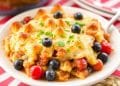 A plate of baked casserole topped with melted cheese, garnished with fresh blueberries, cherry tomatoes, and green herbs—perfect for fourth of July casserole recipes—served on a white dish with a red and white striped cloth in the background.