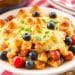 A plate of baked casserole topped with melted cheese, garnished with fresh blueberries, cherry tomatoes, and green herbs—perfect for fourth of July casserole recipes—served on a white dish with a red and white striped cloth in the background.
