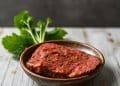 A wooden bowl holds a slice of uncooked, seasoned plant-based meat. Fresh green herbs are placed on the rustic wooden surface in the background, perfect for those exploring free carnivore diet recipes with a modern twist.