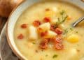 A close-up of a bowl of creamy potato soup recipe with chunks of potato, crispy bacon bits, and garnish, with a spoon in the bowl.
