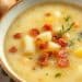 A close-up of a bowl of creamy potato soup recipe with chunks of potato, crispy bacon bits, and garnish, with a spoon in the bowl.