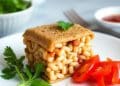 A square slice of baked beans sandwiched between two pieces of bread, garnished with parsley, sits on a white plate with sliced cherry tomatoes—a Quick Recipe from Jalbiteworldfood. Bowls of herbs and sauce are blurred in the background.