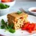 A square slice of baked beans sandwiched between two pieces of bread, garnished with parsley, sits on a white plate with sliced cherry tomatoes—a Quick Recipe from Jalbiteworldfood. Bowls of herbs and sauce are blurred in the background.