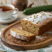 A sliced loaf of cooks zucchini bread dusted with powdered sugar sits on a wooden tray, with a cup of tea and a cucumber in the background.