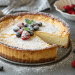 A baked ricotta cheesecake with a golden crust, topped with powdered sugar, blueberries, raspberries, and a sprig of mint. One slice is cut and slightly pulled out. A bowl of berries and a checkered napkin are in the background.