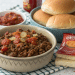 A bowl of seasoned ground beef with tomatoes and onions sits next to sandwich buns, a packet of cheese sauce, and salsa—perfect for an easy sloppy joe recipe with just 3 ingredients on a checkered cloth.