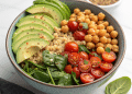 A high protein vegan salad bowl filled with sliced avocado, cooked chickpeas, halved cherry tomatoes, fresh spinach leaves, and quinoa arranged in sections. A small dish with seeds is visible in the background.