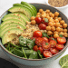 A high protein vegan salad bowl filled with sliced avocado, cooked chickpeas, halved cherry tomatoes, fresh spinach leaves, and quinoa arranged in sections. A small dish with seeds is visible in the background.