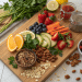 A wooden board with sliced oranges, cucumbers, strawberries, blueberries, papaya, nuts, seeds, parsley, whole grain crackers, dried cranberries—perfect inspiration for raw vegan recipes. Fresh herbs and lemon are nearby with jars of salsa and chia seeds.