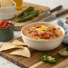 A bowl of cheesy rotel dip recipe, topped with diced tomatoes and green onions, sits on a wooden board with tortilla chips, sliced jalapeño, lime, and a bowl of salsa nearby.