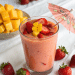 A glass of Strawberry Mango Smoothie topped with sliced strawberries, mango cubes, a yellow flower, and a decorative paper umbrella, surrounded by fresh strawberries and a bowl of mango cubes on a white surface.