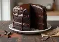 A three-layer chocolate cake with glossy chocolate ganache dripping down the sides, sitting on a white plate. A large slice is missing, revealing the rich, moist interior. A fork and chocolate shavings are nearby on a wooden table.
