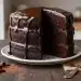 A three-layer chocolate cake with glossy chocolate ganache dripping down the sides, sitting on a white plate. A large slice is missing, revealing the rich, moist interior. A fork and chocolate shavings are nearby on a wooden table.