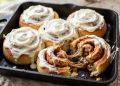 Pan of golden cinnamon rolls with cream cheese frosting, one roll pulled apart showing the gooey swirl