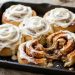Pan of golden cinnamon rolls with cream cheese frosting, one roll pulled apart showing the gooey swirl