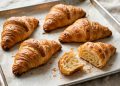 Golden-brown flaky croissants with visible layers on parchment, one broken open showing airy interior