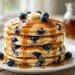 Tall stack of fluffy blueberry buttermilk pancakes with maple syrup dripping and fresh blueberries