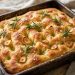 Golden focaccia with deep dimples, fresh rosemary, roasted garlic slices, and flaky salt in a pan