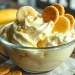 A glass bowl filled with creamy banana pudding, topped with banana slices and vanilla wafers, sits on a wooden table beside bananas—perfect inspiration for Banana Pudding Ice Cream.