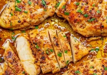 Close-up of juicy, golden-brown Garlic Butter Chicken breasts sprinkled with herbs and spices, one piece sliced to reveal the moist interior, all surrounded by a rich, savory sauce.
