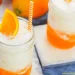 Two mason jars filled with a creamy, orange Dirty Soda, topped with ice, an orange slice, and an orange-striped straw, sit on light coasters with a blue cloth underneath.