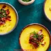 Three bowls of creamy yellow Fall Soup topped with chopped bacon and fresh chives, arranged on a teal surface. A small bowl of chopped chives sits nearby.