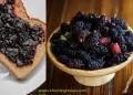 A slice of bread with dark berry jam, possibly made from mulberries, sits next to a bowl filled with fresh blackberries and a small jar of jam on a wooden surface.