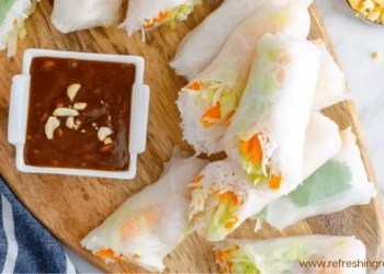 A wooden board with fresh Summer Rolls filled with vegetables and rice noodles, sliced in half, served with a small dish of brown dipping sauce topped with chopped peanuts. A blue and white towel is partially visible on the side.