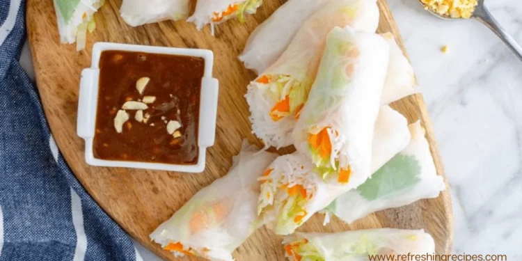 A wooden board with fresh Summer Rolls filled with vegetables and rice noodles, sliced in half, served with a small dish of brown dipping sauce topped with chopped peanuts. A blue and white towel is partially visible on the side.