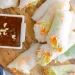 A wooden board with fresh Summer Rolls filled with vegetables and rice noodles, sliced in half, served with a small dish of brown dipping sauce topped with chopped peanuts. A blue and white towel is partially visible on the side.