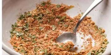 A close-up of a white bowl containing a flavorful meatloaf seasoning recipe—a blend of dry herbs and spices—with a silver spoon partially submerged, ready to elevate your next dish.