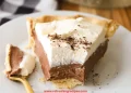 A slice of Chocolate Cream Pie with a flaky crust, creamy chocolate filling, and a thick layer of whipped cream sits on a white plate, a forkful already enjoyed.