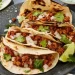 Three tacos filled with seasoned turkey meat, chopped onions, and cilantro are arranged on a gray plate with charred tortillas. This Turkey Tacos Recipe is garnished with lime wedges and fresh cilantro for a burst of flavor.