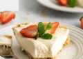 A slice of creamy 3 ingredient no-bake cheesecake garnished with two strawberry halves and a sprig of mint sits on a white plate with a fork. More strawberries and another plate are visible in the background.