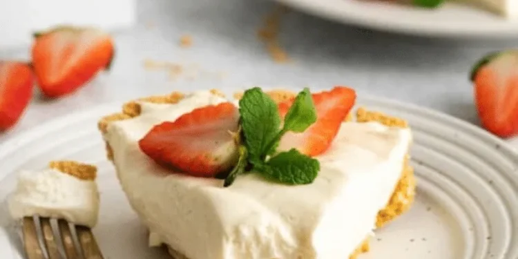 A slice of creamy 3 ingredient no-bake cheesecake garnished with two strawberry halves and a sprig of mint sits on a white plate with a fork. More strawberries and another plate are visible in the background.