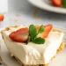 A slice of creamy 3 ingredient no-bake cheesecake garnished with two strawberry halves and a sprig of mint sits on a white plate with a fork. More strawberries and another plate are visible in the background.