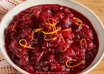 A white bowl filled with chunky cranberry sauce, made from a classic Cranberry Sauce Recipe and garnished with thin strips of orange zest, sits on a wooden surface with a patterned cloth nearby.