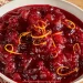A white bowl filled with chunky cranberry sauce, made from a classic Cranberry Sauce Recipe and garnished with thin strips of orange zest, sits on a wooden surface with a patterned cloth nearby.