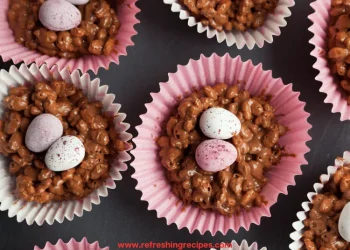 Rice Krispie Buns in pink paper cases, each topped with two speckled candy eggs, are arranged on a dark surface. The website refreshingrecipes.com is visible at the bottom.
