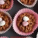 Rice Krispie Buns in pink paper cases, each topped with two speckled candy eggs, are arranged on a dark surface. The website refreshingrecipes.com is visible at the bottom.