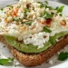 A slice of whole grain toast topped with mashed avocado, cottage cheese, a cooked egg, crumbled cheese, chopped parsley, and sprinkled spices—an easy addition to your favorite cottage cheese recipes, served on a white plate.